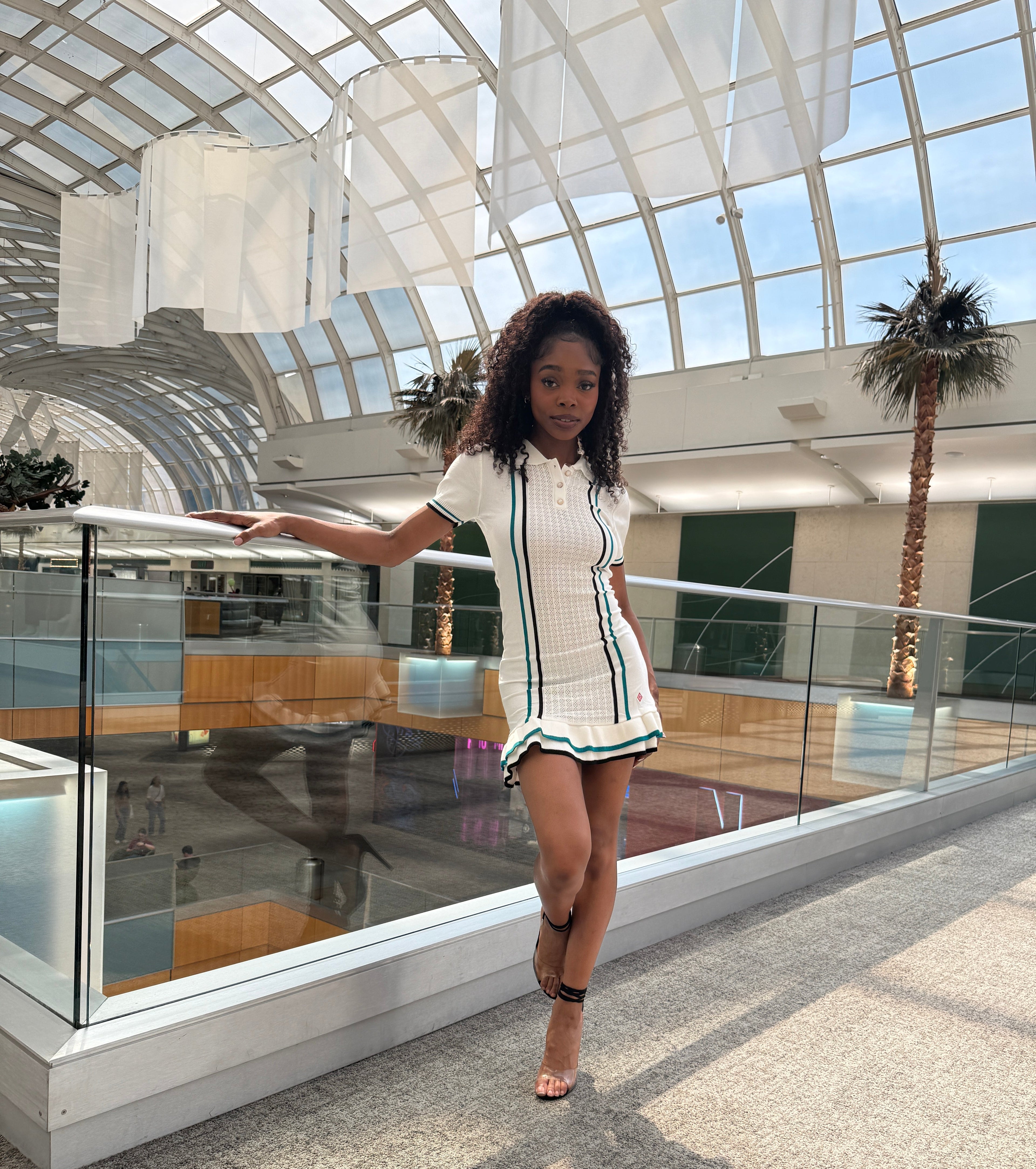 Model in our Striped Tennis dress standing in a modern indoor setting with glass walls and palm trees.